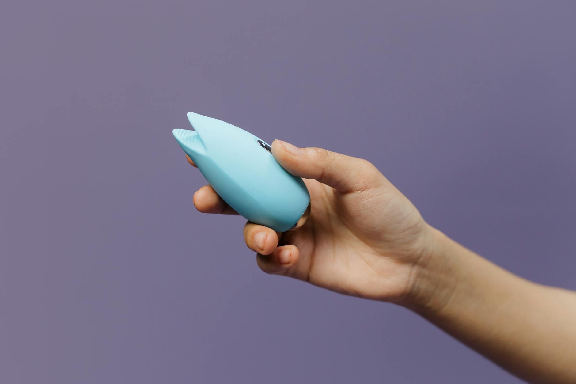 A blue silicone clitoral vibrator held in hand against a solid purple background, promoting self-love and sexuality.