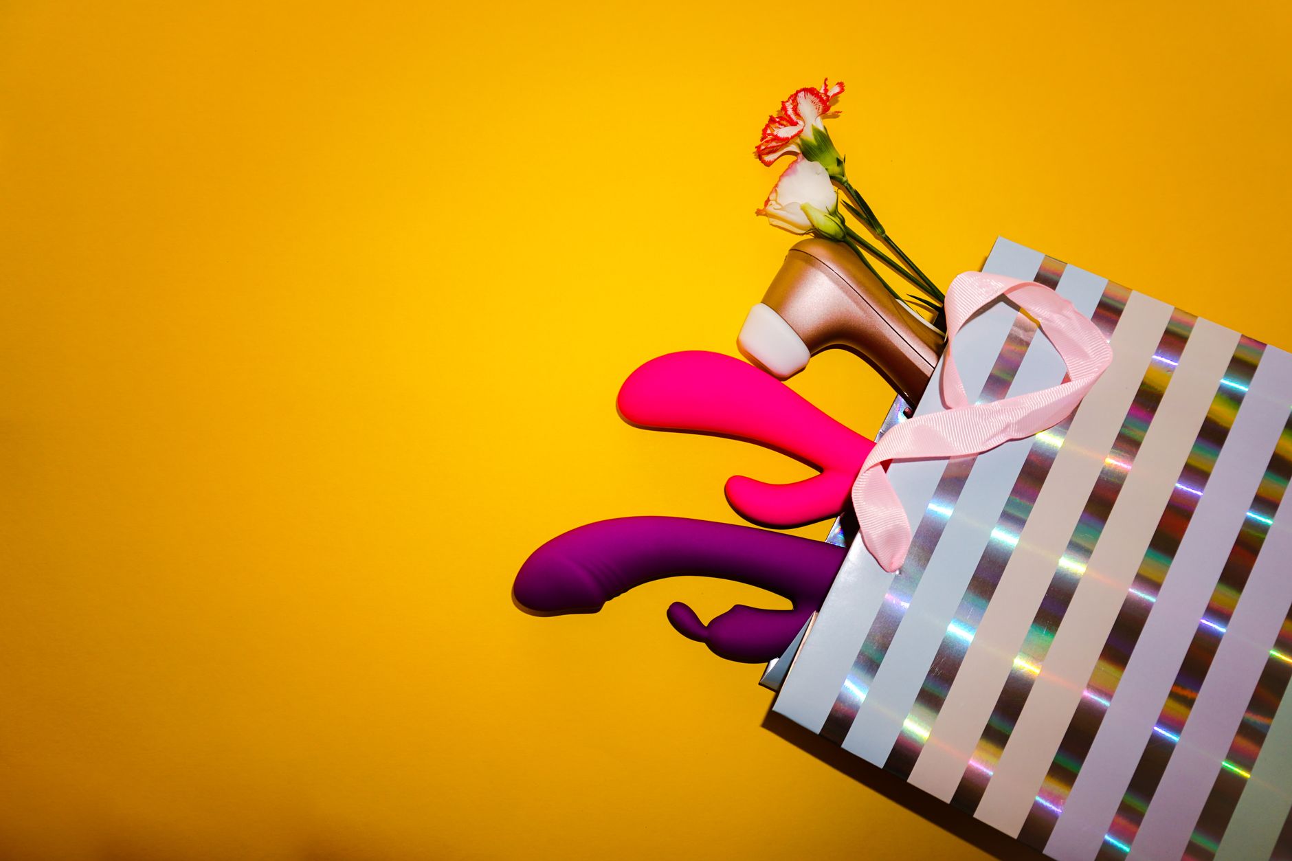Vibradores coloridos com flores em uma bolsa de presente holográfica em fundo amarelo vibrante
